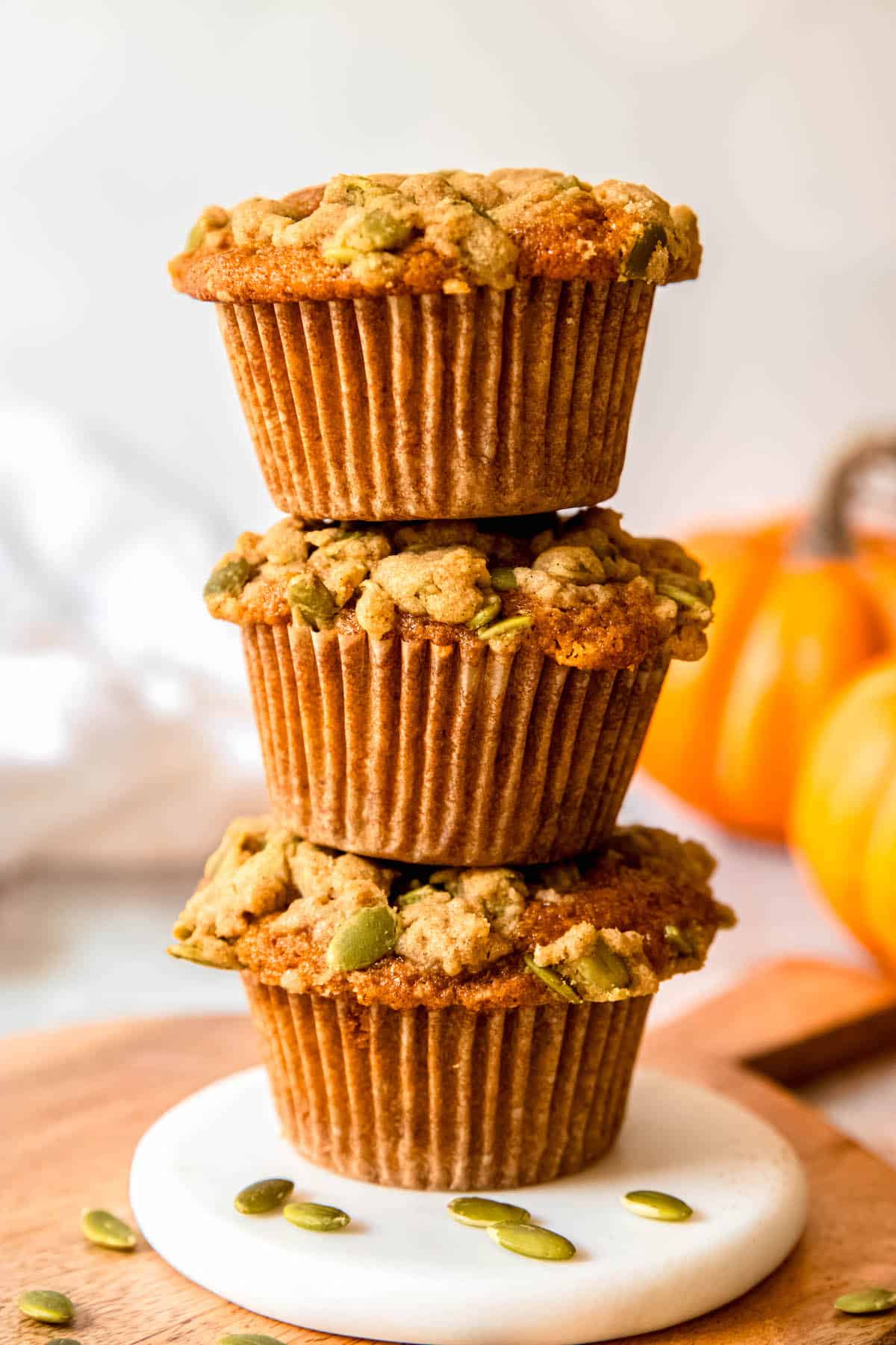 stack of three banana and pumpkin muffins on a white coaster; the pumpkin spice streusel topping is causing them to tilt left and right for a slightly wonky stack.