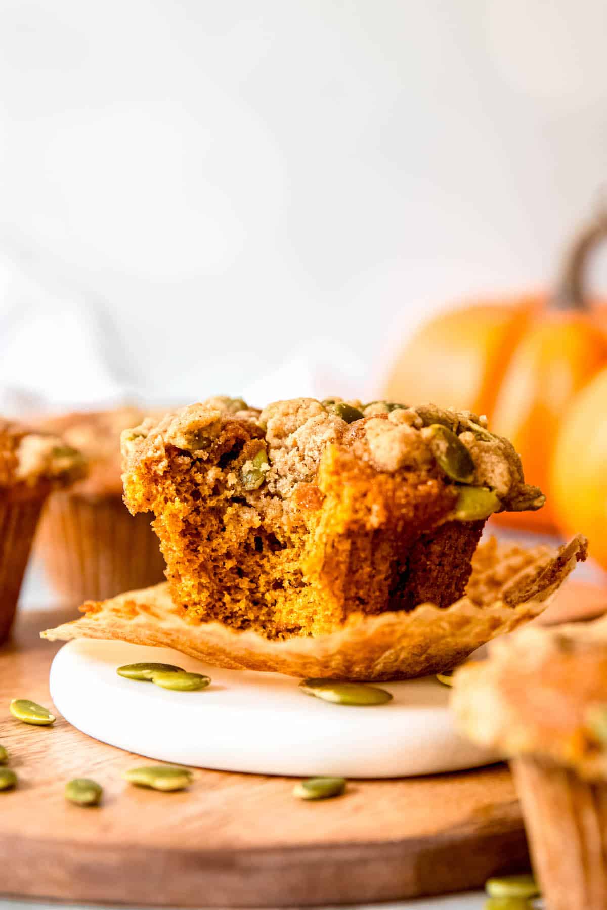 side on shot of a banana pumpkin streusel muffin with the paper liner pulled down and a bite taken out on top of a white coaster on a wooden serving tray with pumpkin seeds in the foreground and pumpkins in the background.