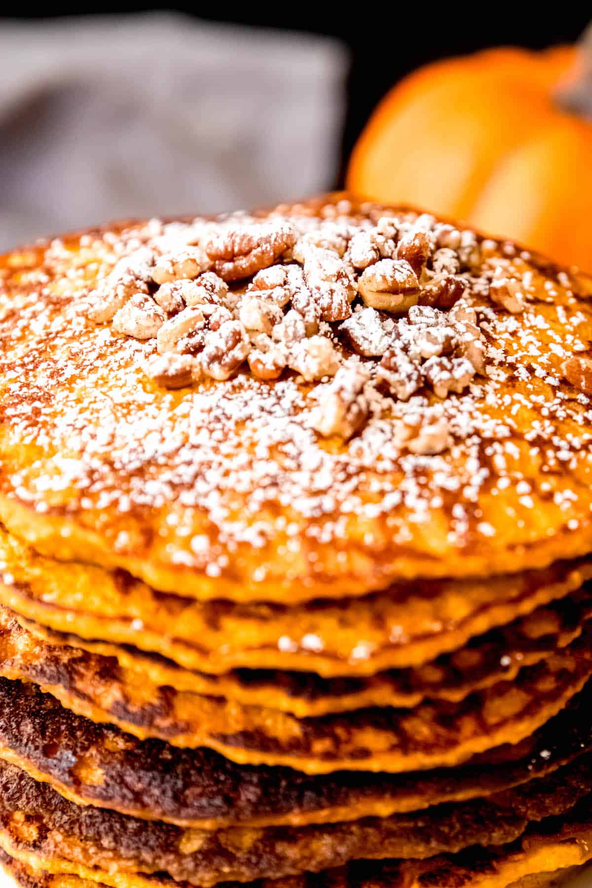 closeup shot of the top of a stack of blender oat pumpkin protein pancakes after topping with chopped pecans and powdered sugar with a mini pumpkin and a grey cloth blurred out in the background.