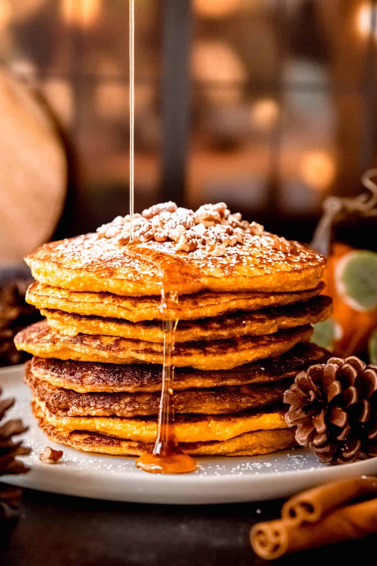 action shot of maple syrup being drizzled atop a stack of healthy pumpkin protein pancakes topped with pecans and powdered sugar.