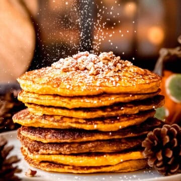 action shot of cottage cheese pumpkin protein pancakes being dusted with powdered sugar in front of a cozy fairy-lit window.