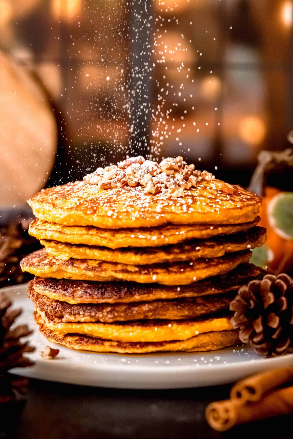 action shot of cottage cheese pumpkin protein pancakes being dusted with powdered sugar in front of a cozy fairy-lit window.