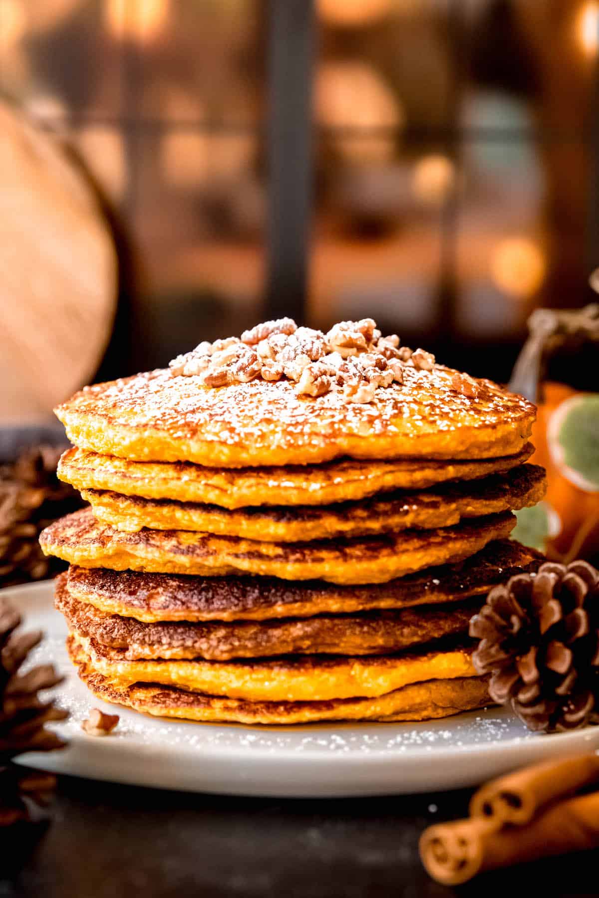 stack of 8 pumpkin protein pancakes made with oats and cottage cheese on a white plate on a dark table with cinnamon sticks and pine cones scattered around.