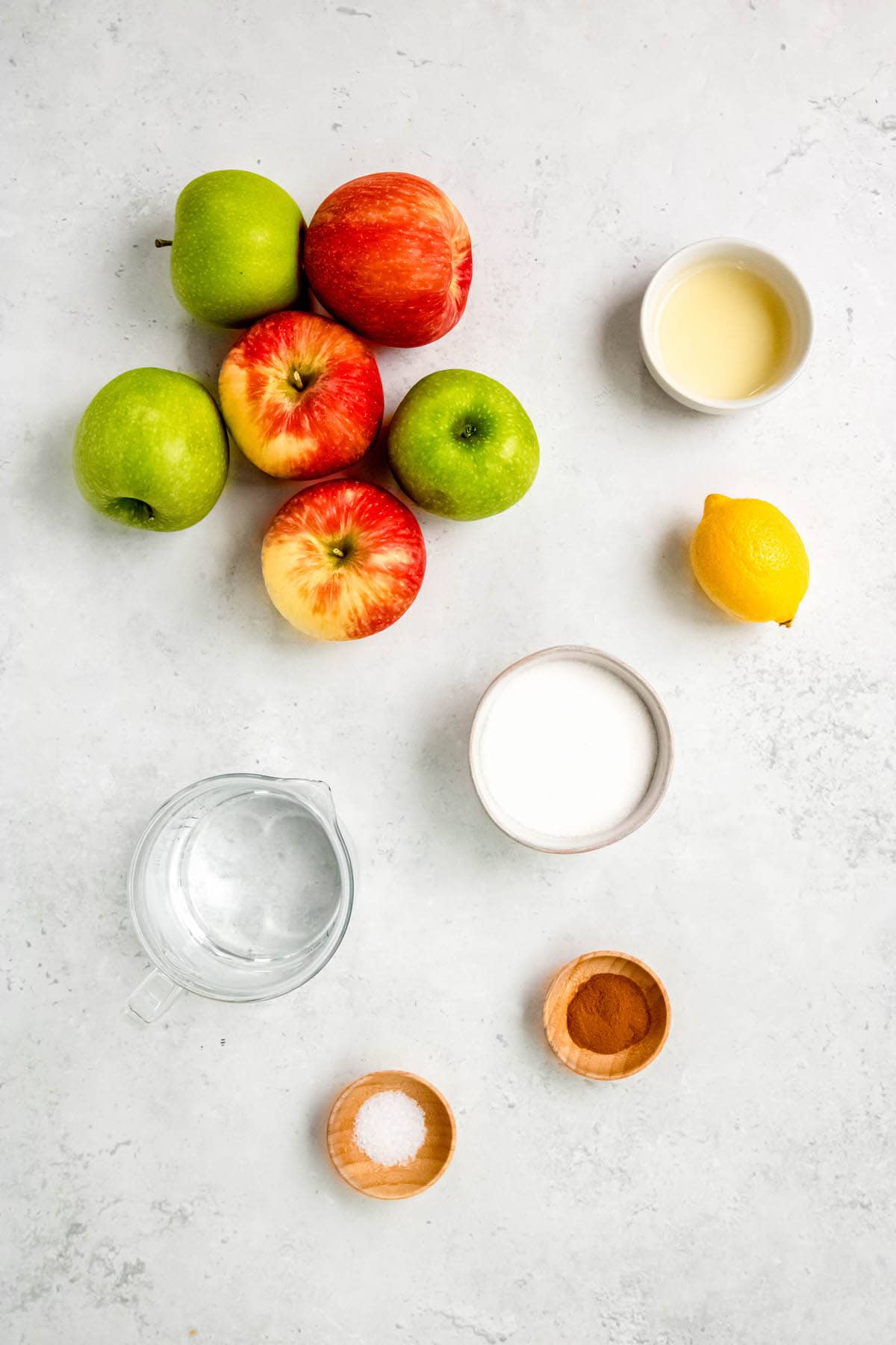 fresh apples, apple cider vinegar, a whole lemon, sugar, cinnamon, salt, and water measured out into bowls on a white table.