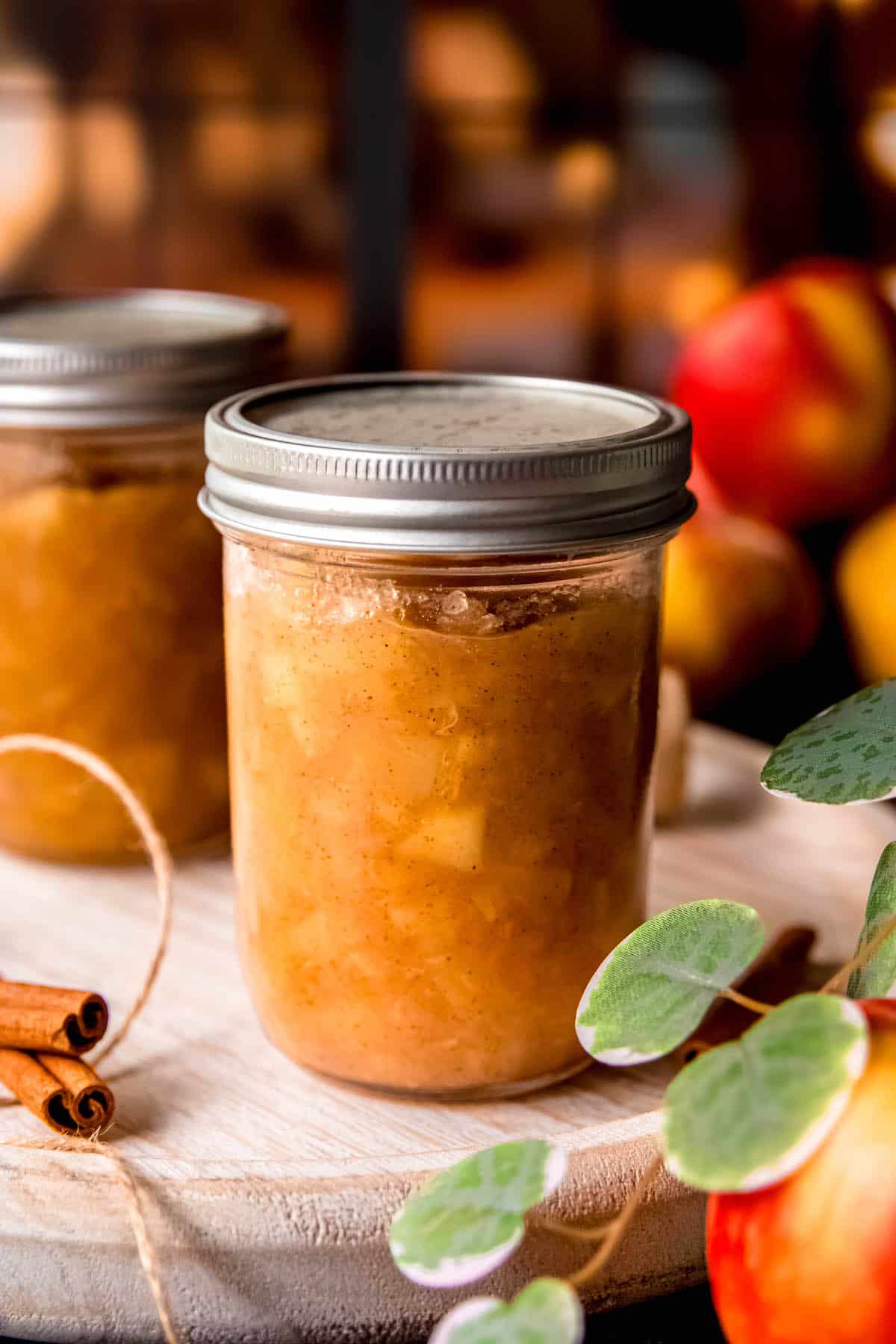 two canned mason jars of homemade spiced apple preserves on a wooden tray with a fairy-lit black-paned window behind.