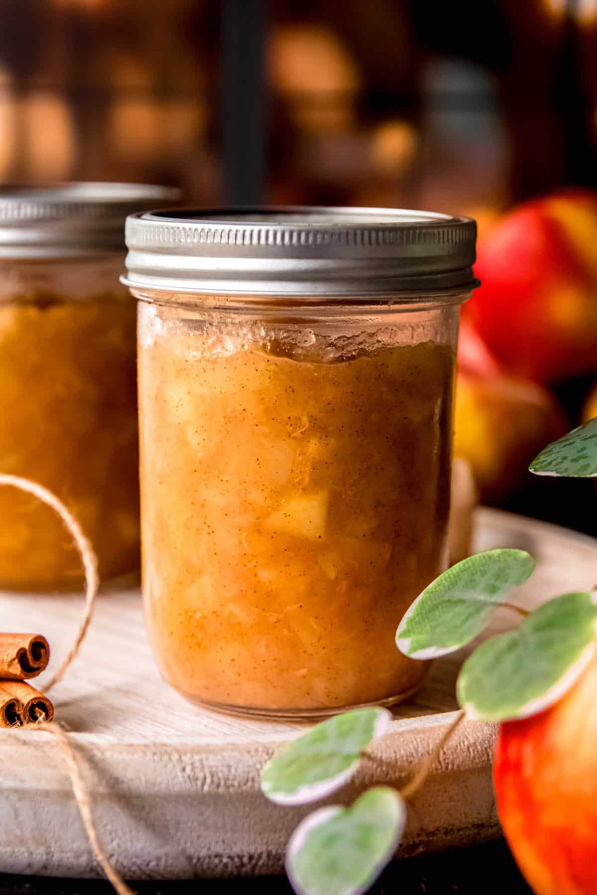 2 mason jars of cinnamon apple jam after canning in a water bath with whole apples blurred out in the background, cinnamon sticks and whole apple in the foreground.