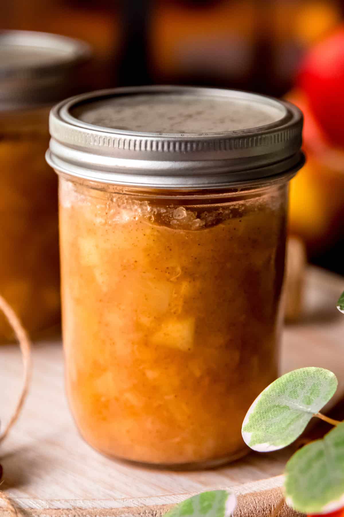 side-on shot of a can of apple preserves that can be used as applesauce, apple jam, or apple pie filling on a wooden tray with another full jar and whole apples behind and leaves from a green houseplant.