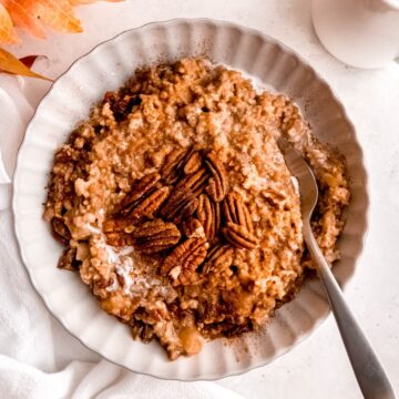 square flat lay shot of a bowl of cinnamon apple steel cut oats topped with a handful of pecans and a drizzle of cream on a white table with a few fall leaves.