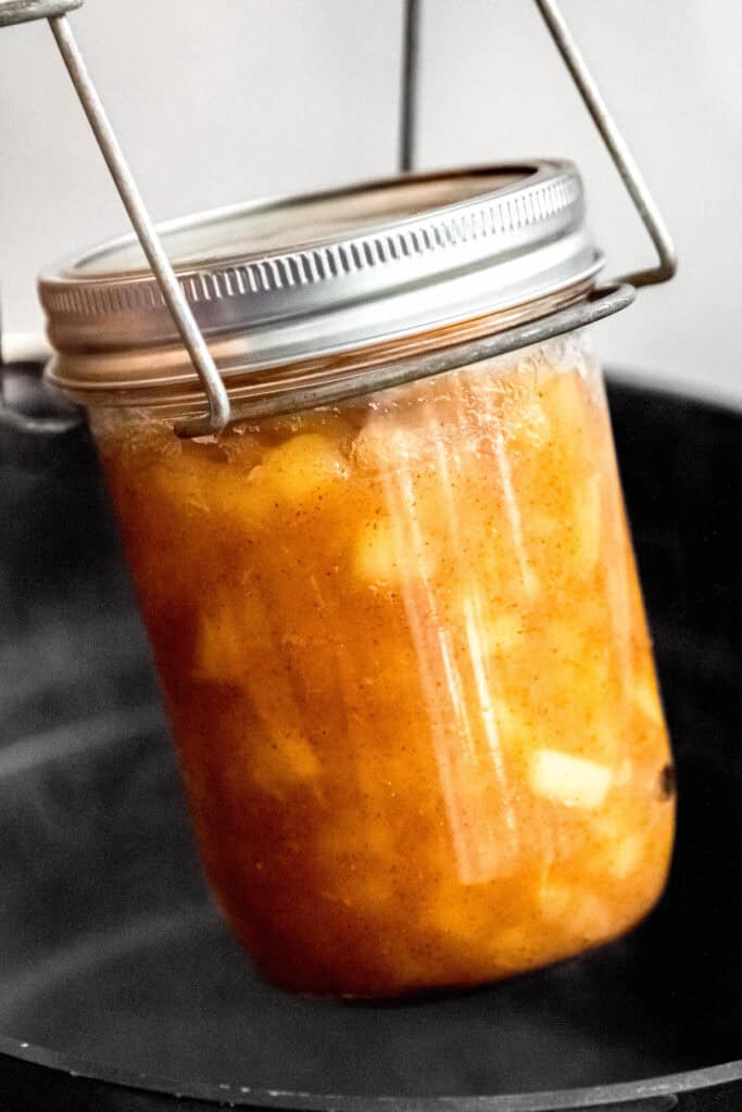 mason jar of apple preserves being lifted out of a water bath using canning tongs after boiling for shelf-stable preservation.