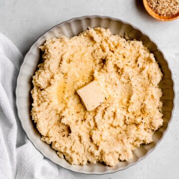 square hero image of keto garlic roasted cauliflower mash in a grey scalloped serving bowl on a white table with a wooden pinch bowl of garlic parmesan seasoning.