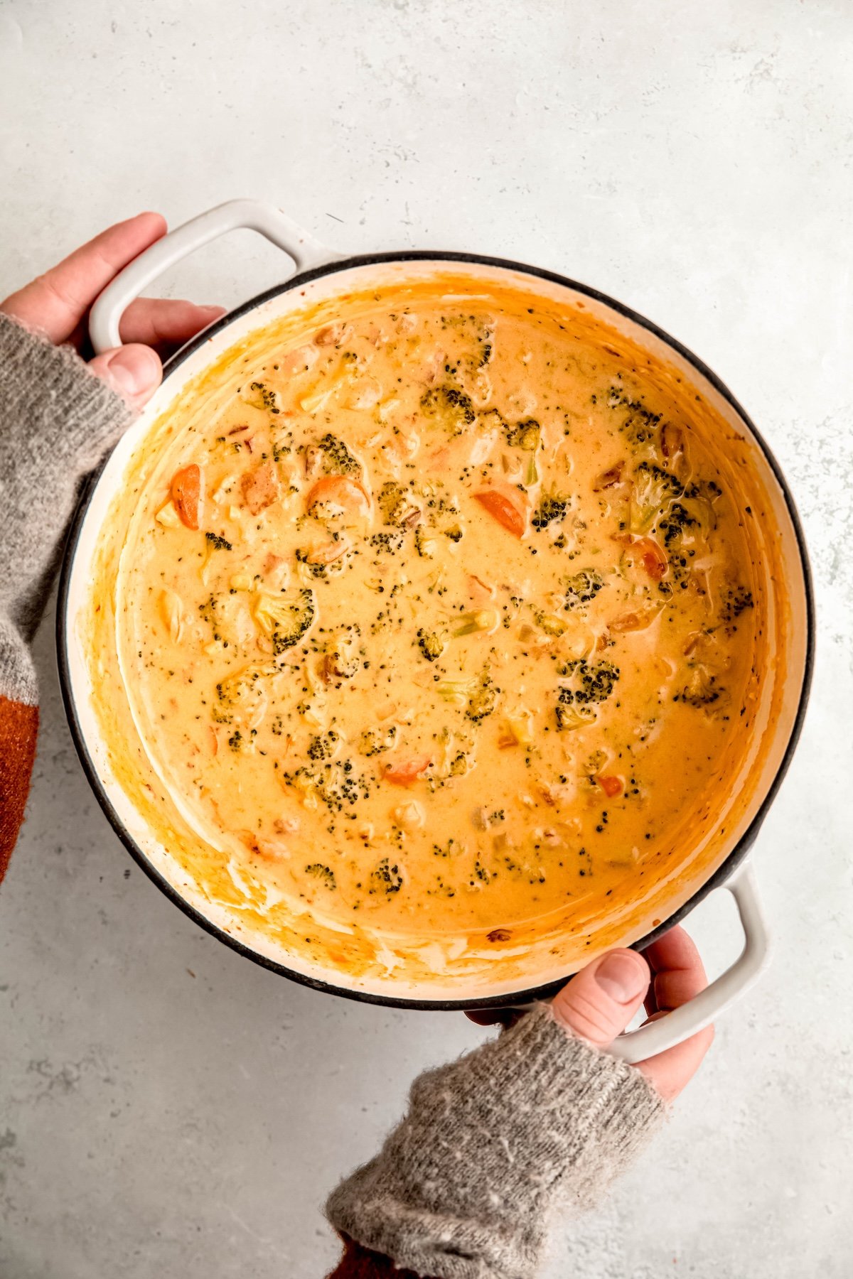 hands grabbing the handles of a white enameled dutch oven filled with bacon broccoli cheddar cheese soup from a white table.