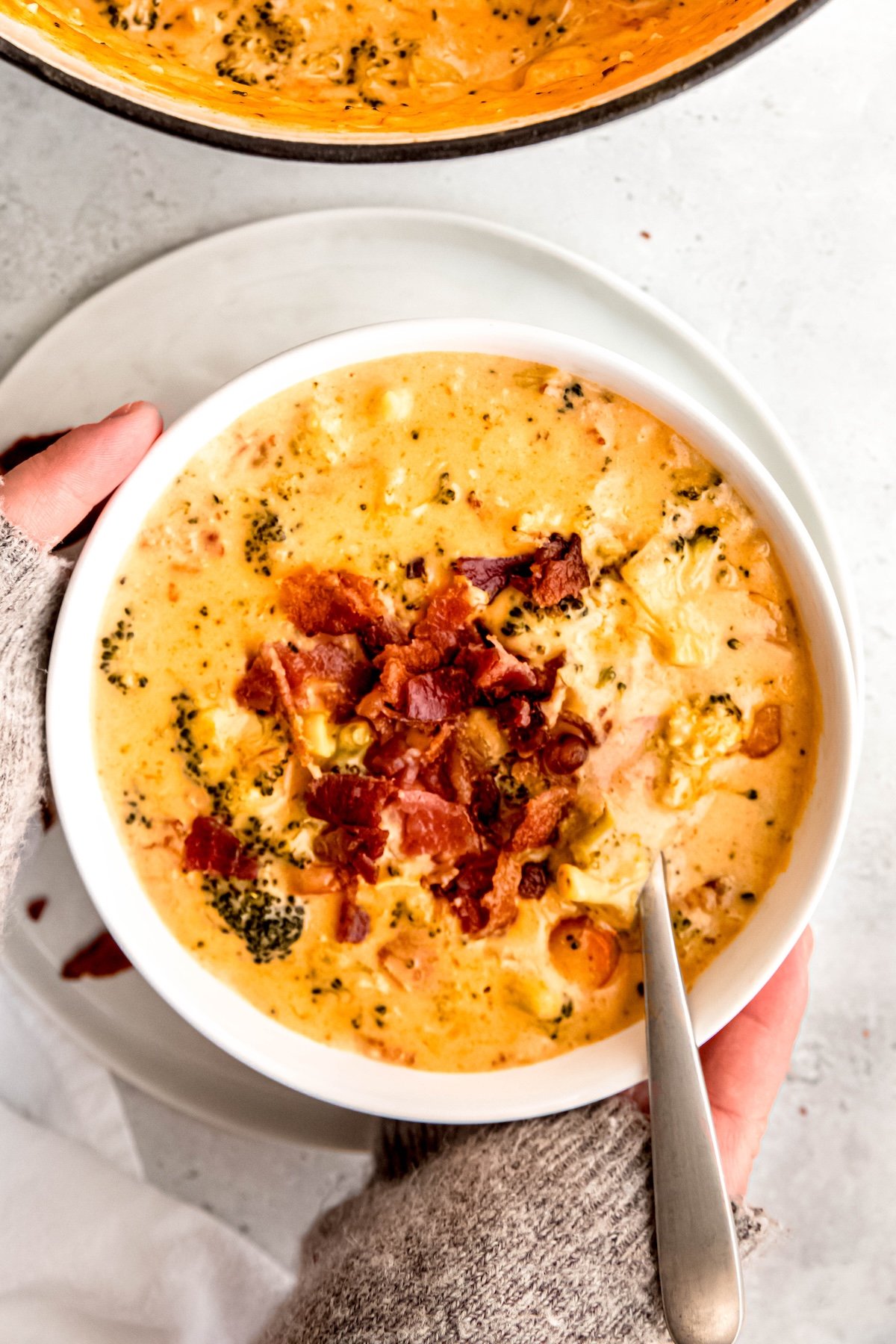 two hands in grey sweater pulled down to the knuckles grabbing a bowl of bacon broccoli cheese soup with the rest of the pot of it in the top of the frame.
