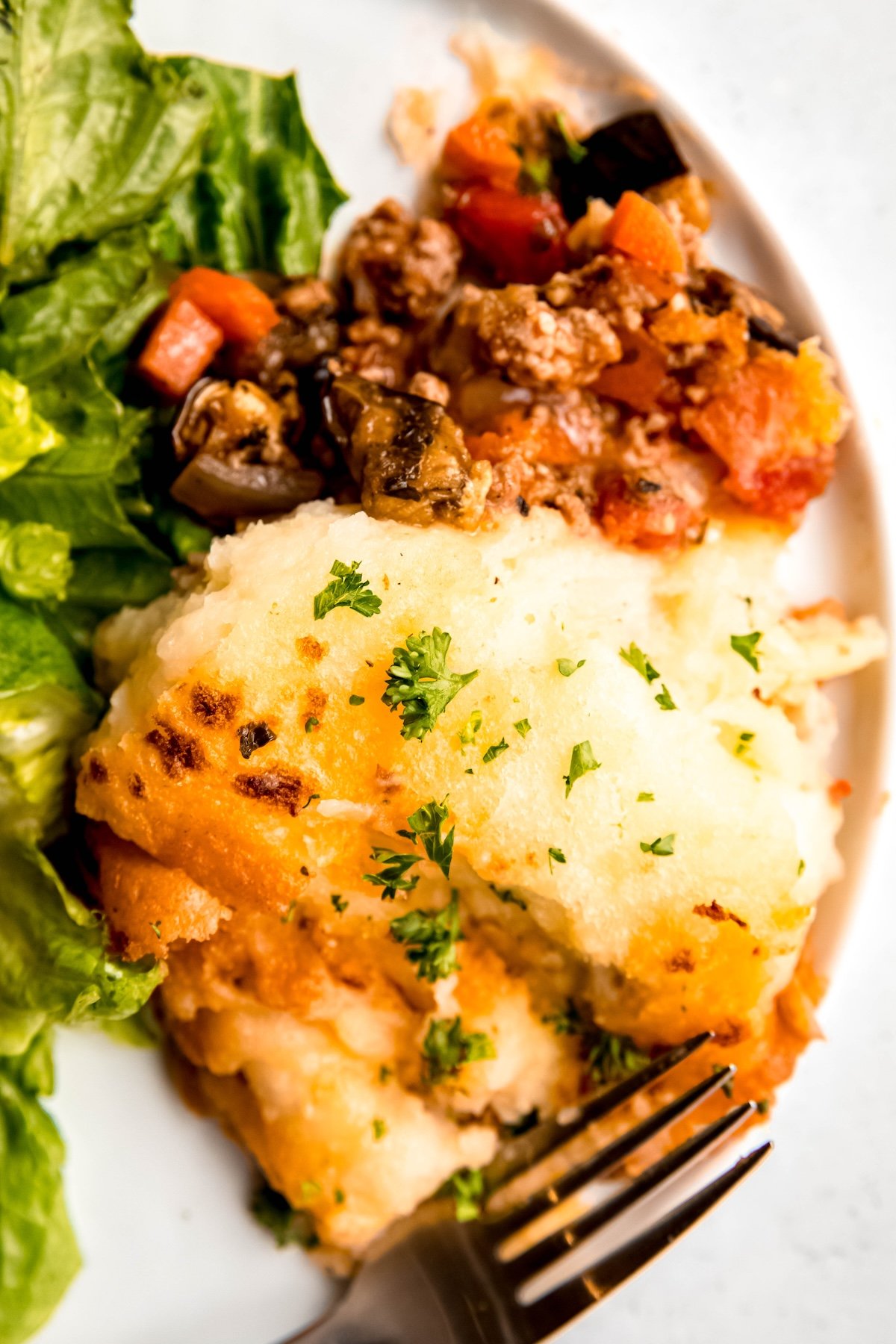 closeup shot of a serving of GF shepherd's pie on half of a white round dinner plate with the head of a fork in the bottom of the screen and a sliver of green from the side salad on the left of the frame.