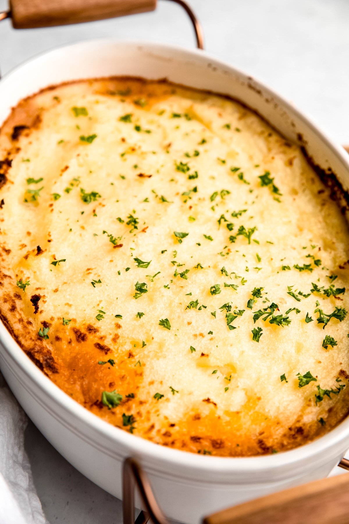 45 degree angle shot of an eggplant ground lamb gluten free shepherd's pie in an oval white casserole dish with metal and wood serving handles attached; the top of the potato layer is sprinkled with fresh parsley and the edges of the dish have the potatoes turning golden brown and caught.