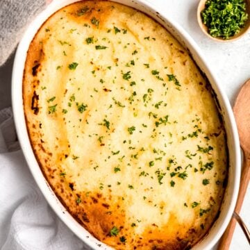 two hands wrapped in a cozy grey sweather to the knuckles grabbing the serving handles of the gluten free moussaka shepherd's pie from the table.
