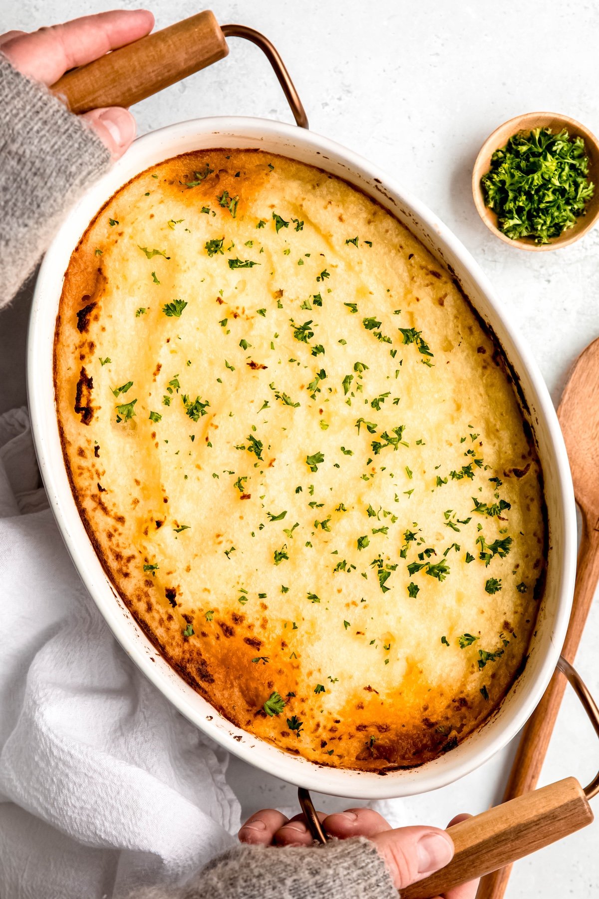 two hands wrapped in a cozy grey sweather to the knuckles grabbing the serving handles of the gluten free moussaka shepherd's pie from the table.