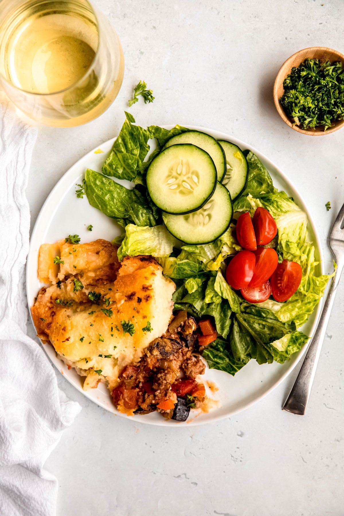 overhead shot of a dinner plate of gluten-free shepherd's pie made with ground lamb and eggplant with a side salad with romaine, halved cherry tomatoes, and cucumber slices on a white table with a silver fork, a glass of white wine, and a pinch bowl of chopped parsley.