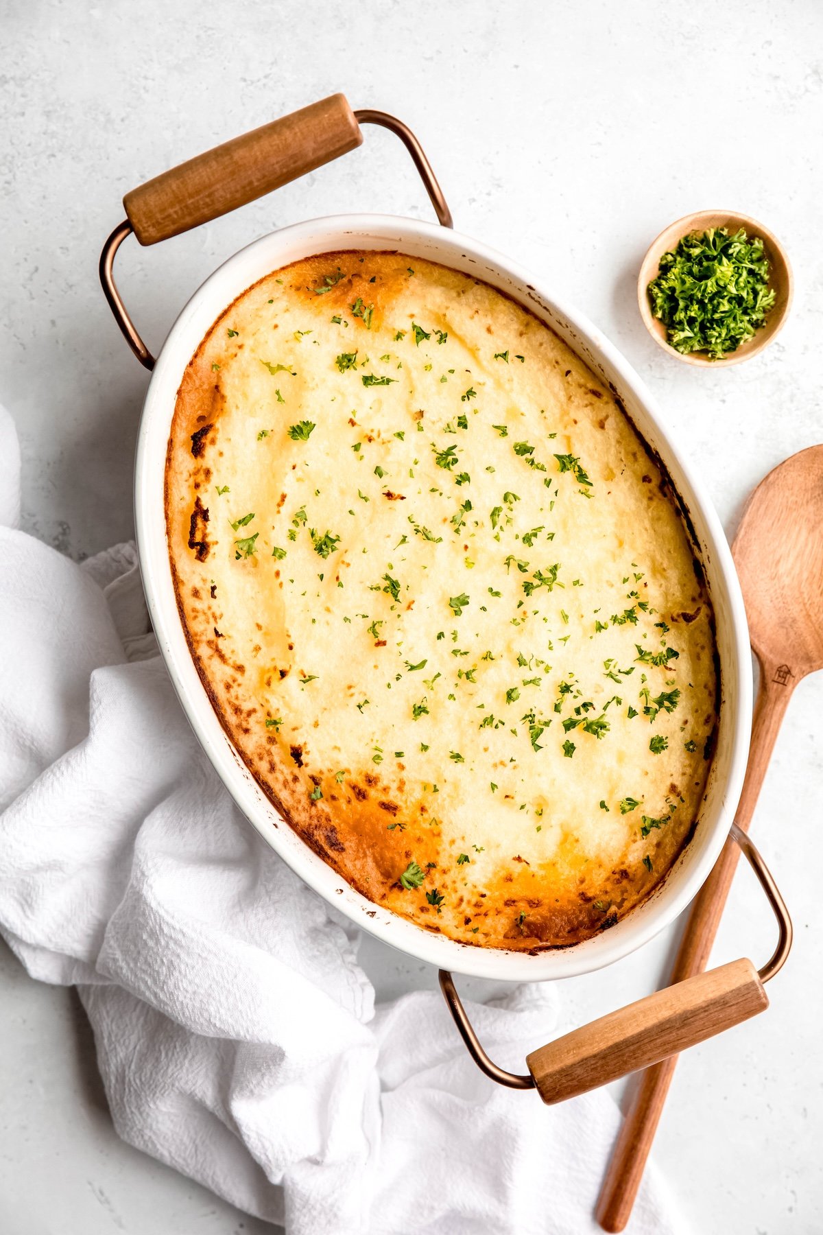 completed Greek gluten free shepherds pie in an oval white casserole dish with wooden and metal handles on a white table with a wooden serving spoon and a small wooden pinch bowl of parsley to garnish to the right and a white linen kitchen towel to the left.