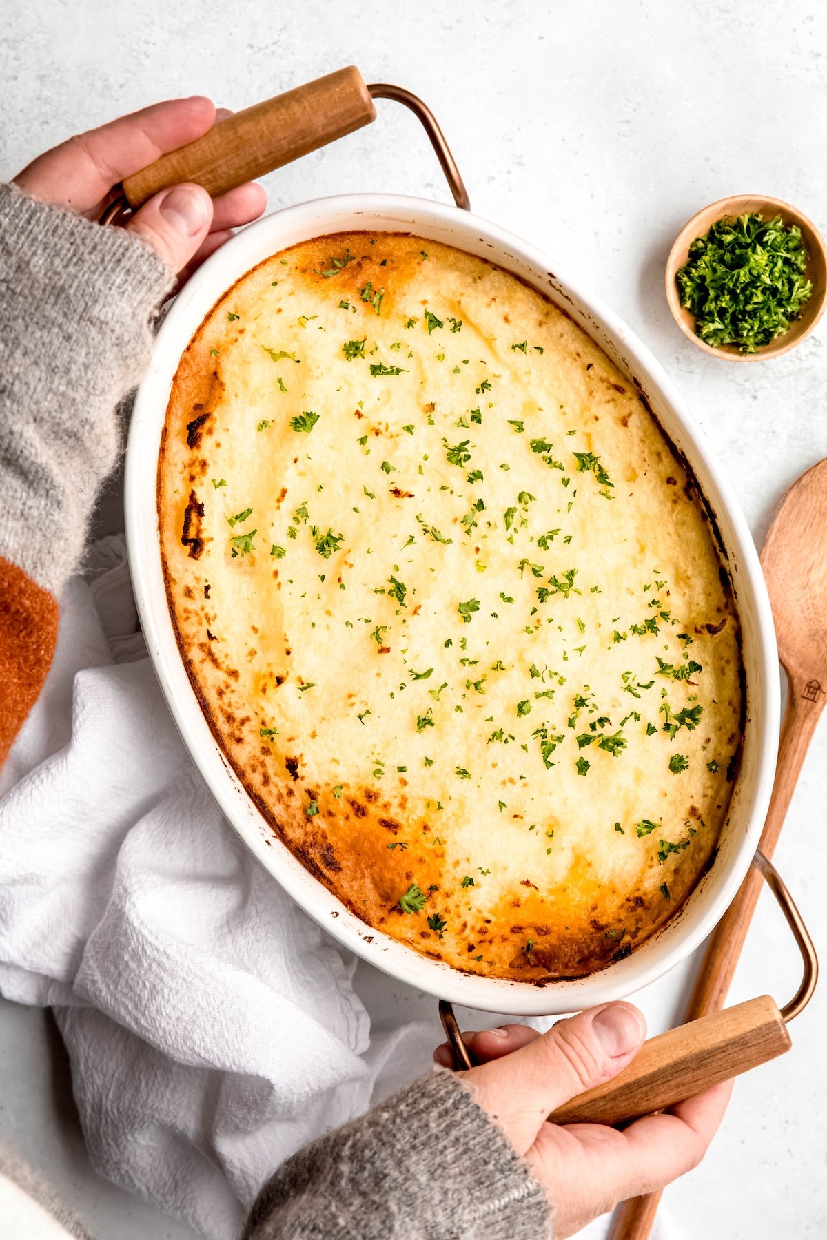 overhead shot of two hands in a cozy grey sweater with brown elbow patches setting down the ground lamb shepherds pie on a white table for serving.