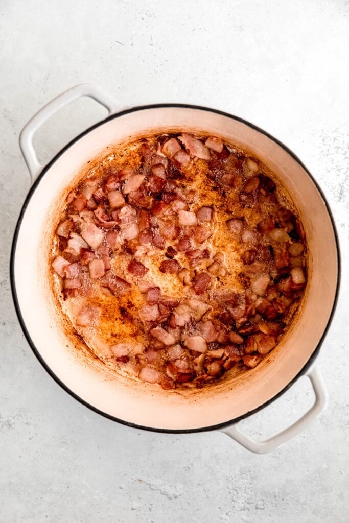 bacon pieces cooking in a white enameled dutch oven; the fat has rendered out and the bacon is becoming darker and crispy.
