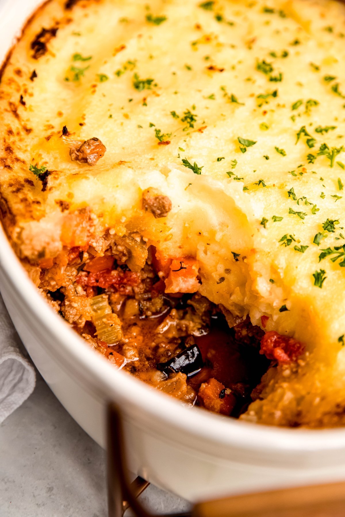 closeup shot of gluten-free greek lamb and eggplant shepherds pie in a casserole dish with a scoop taken out to show the lamb meat sauce, eggplant, and creamy potato layers.