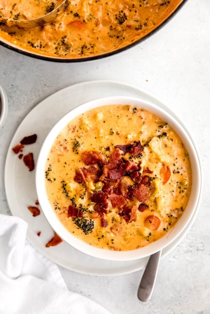 flat lay shot of a white table with a white dinner plate topped with a white bowl of homemade Panera-style bacon cheddar broccoli soup with the rest of the pot in the top ¼ of the frame.