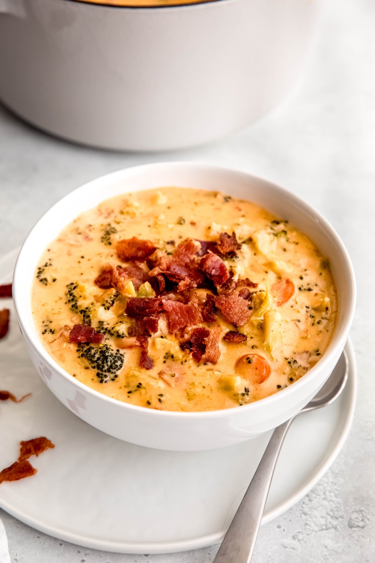 white bowl of panera copycat broccoli cheddar soup with bacon bits on a white dinner plate with a silver spoon.