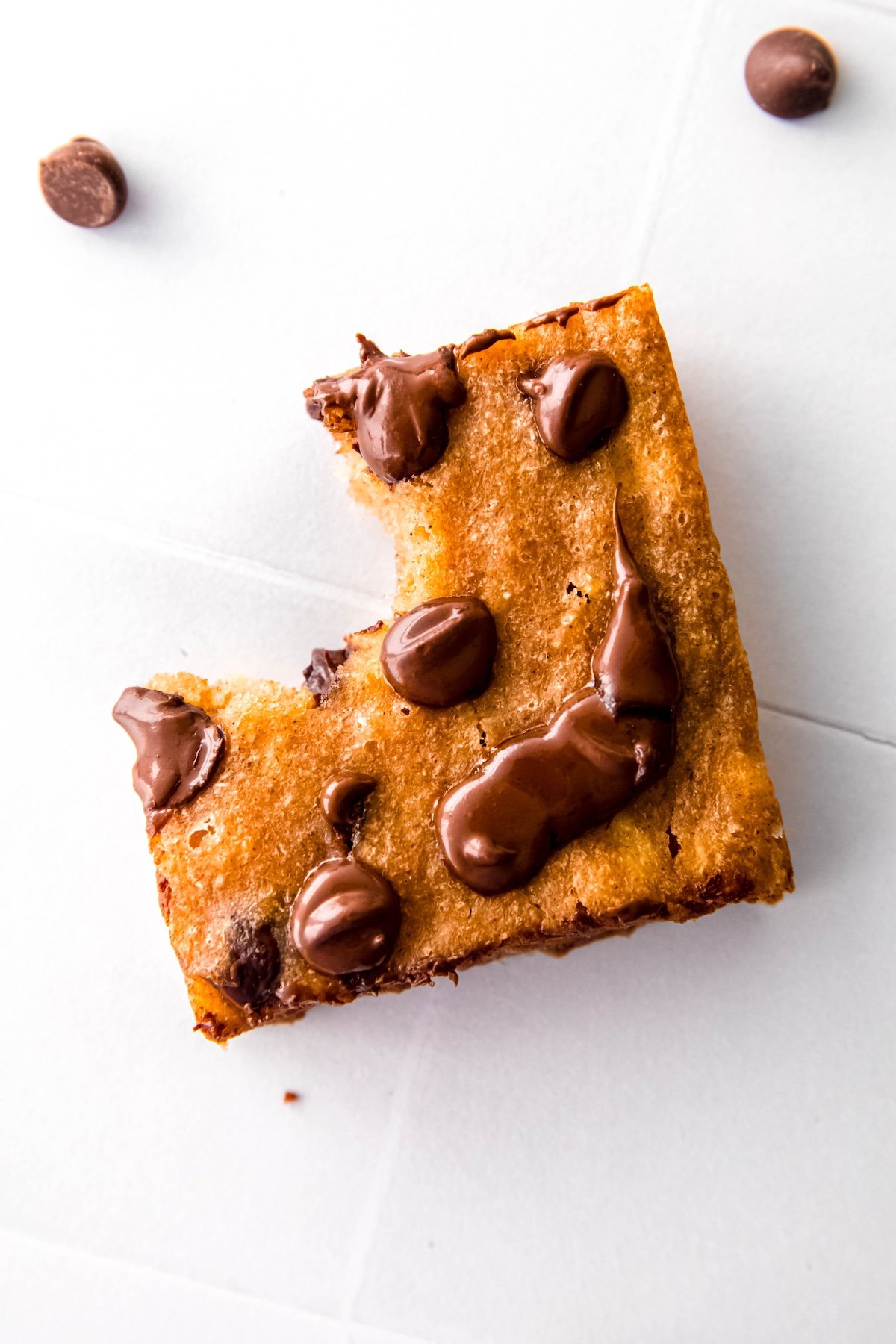 overhead closeup shot of a banana breakfast bar topped with chocolate chips that are all melty on a white table with a few chocolate chips scattered around; a bite has been taken out.