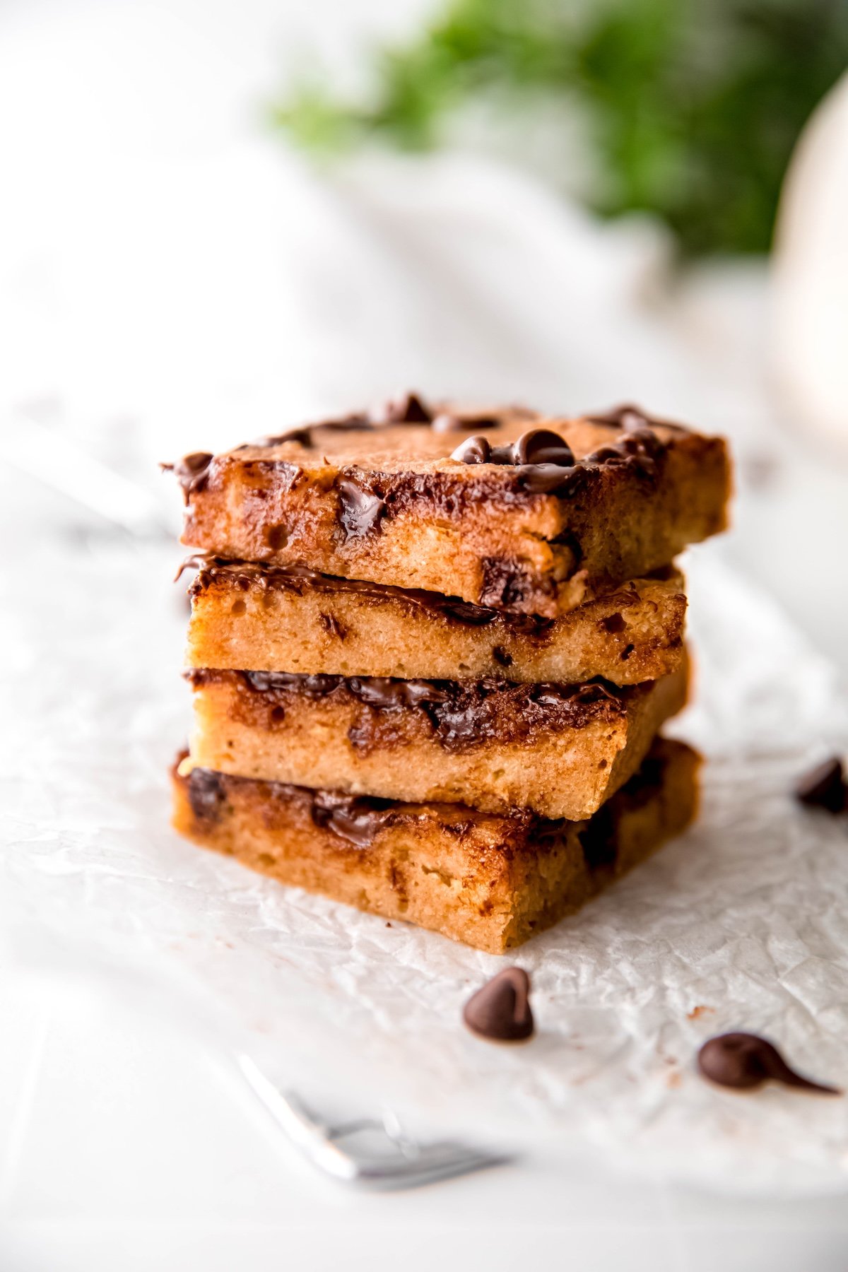 stack of 4 healthy chocolate banana breakfast bars on a white piece of parchment with a few melted chocolate chips strewn around on a white table with a green houseplant blurred out in the background.