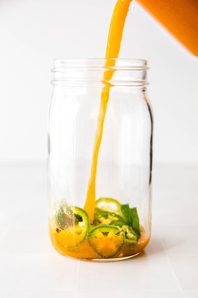 action shot of the turmeric brine being poured into a mason jar with fresh jalapeno slices.