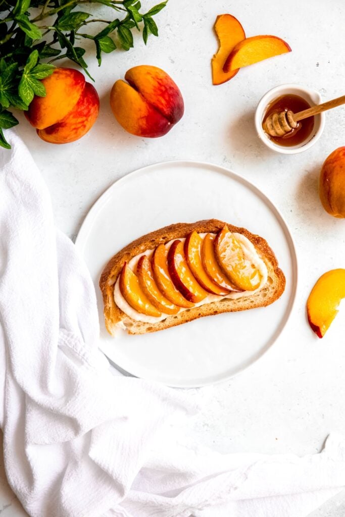 overhead shot of a completed peach breakfast toast after drizzling on the honey on a white table with 2 whole peaches and a green houseplant in the top left corner or the frame, sliced peaches and a ramekin of honey with a wooden honey dipper framing the right side of the plate, and a white linen framing the bottom left corner of the frame.
