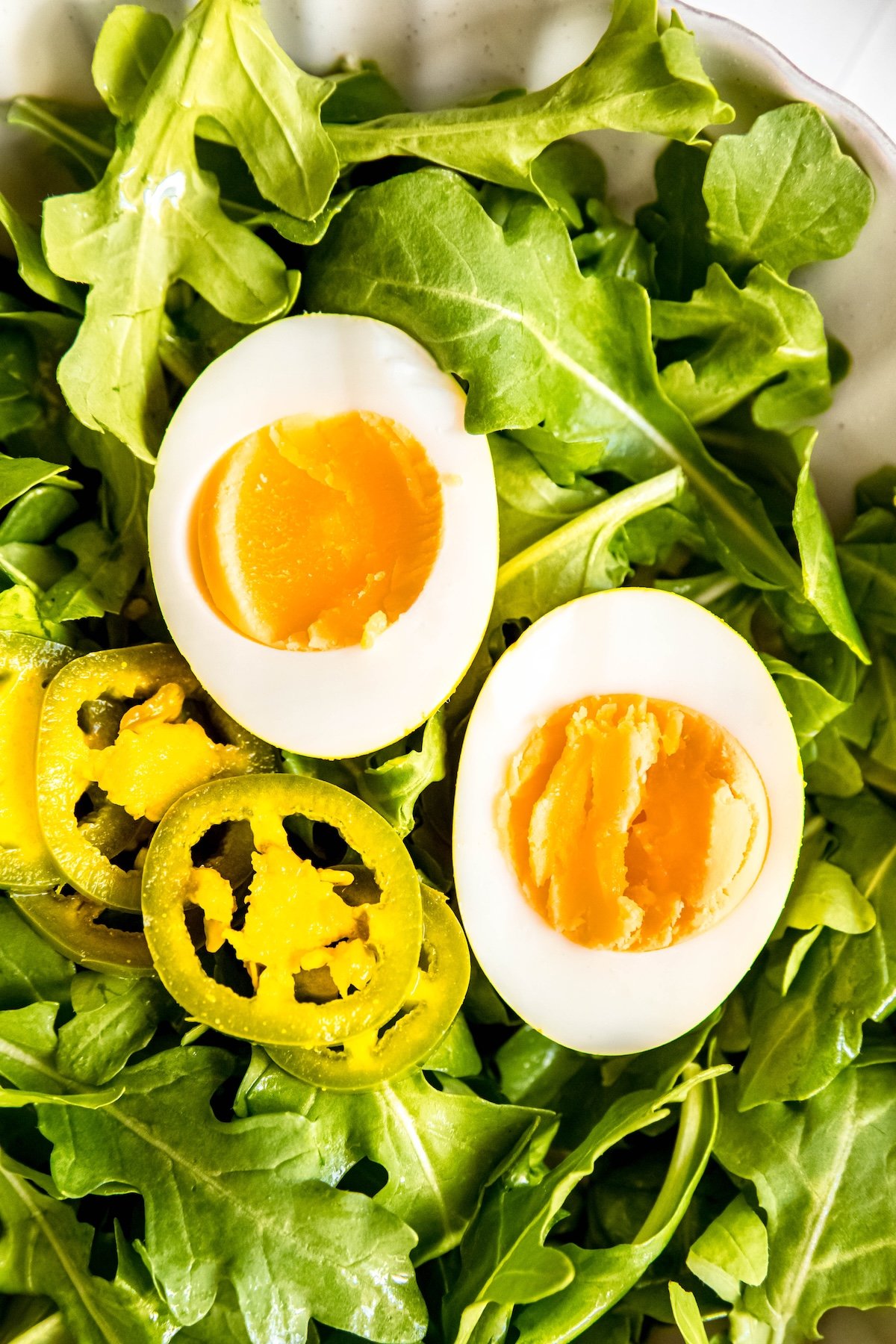 overhead closeup shot of a halved spicy pickled egg on a bed of arugula with slices of the jalapeño it was pickled with; the yolks are bright yellow and soft.