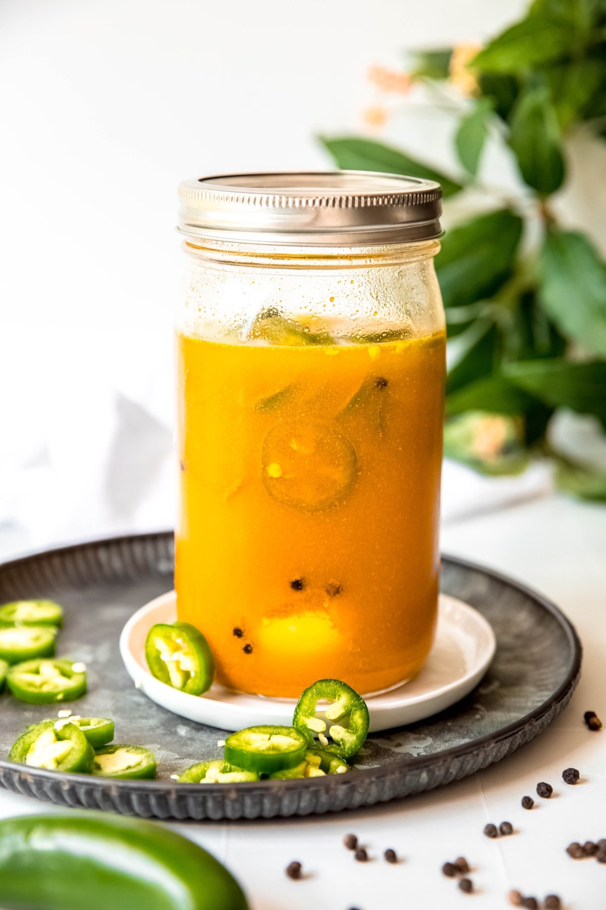 quart-sized mason jar of jalapeño pickled eggs on a white coaster on a hammered black metal serving plate with fresh slices of jalapenos and black peppercorns scattered around.