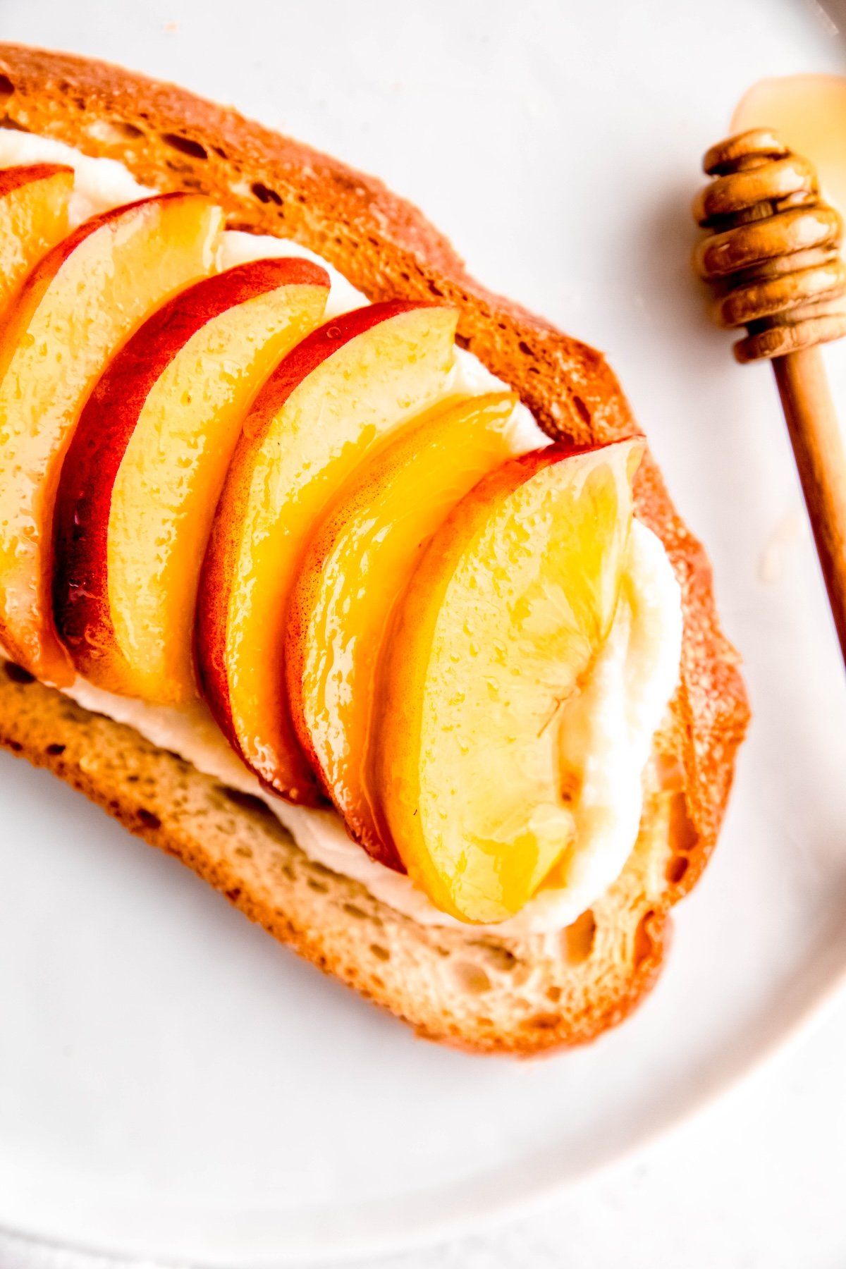 closeup overhead shot of the right half of a peach and ricotta toast on a white plate with a pine-colored wooden honey dipper dripping a puddle of honey on the plate.