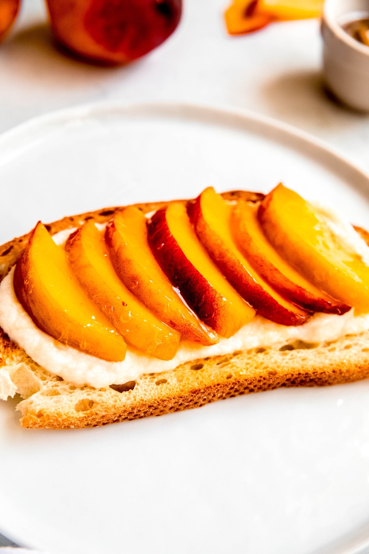 45 degree side on view of ricotta peach toast on a round white plate with 2 fresh whole peaches and a bowl of honey blurred out in the top of the frame.