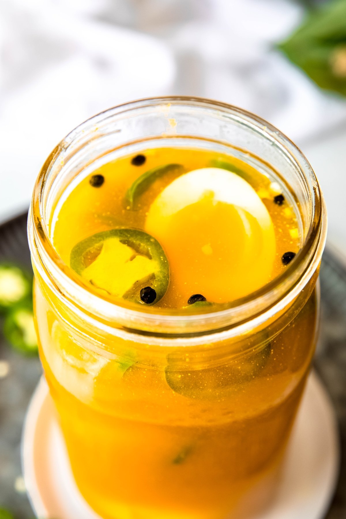 45 degree downward shot into a jar of pickled jalapeno eggs with the top one sliced in half to show the white in contrast to the yellow turmeric brine; there are floating peppercorns and jalapeño slices visible.
