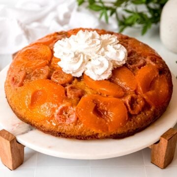square hero image of apricot almond upside down banana cake topped with whipped cream on a white marble and wooden cake stand.