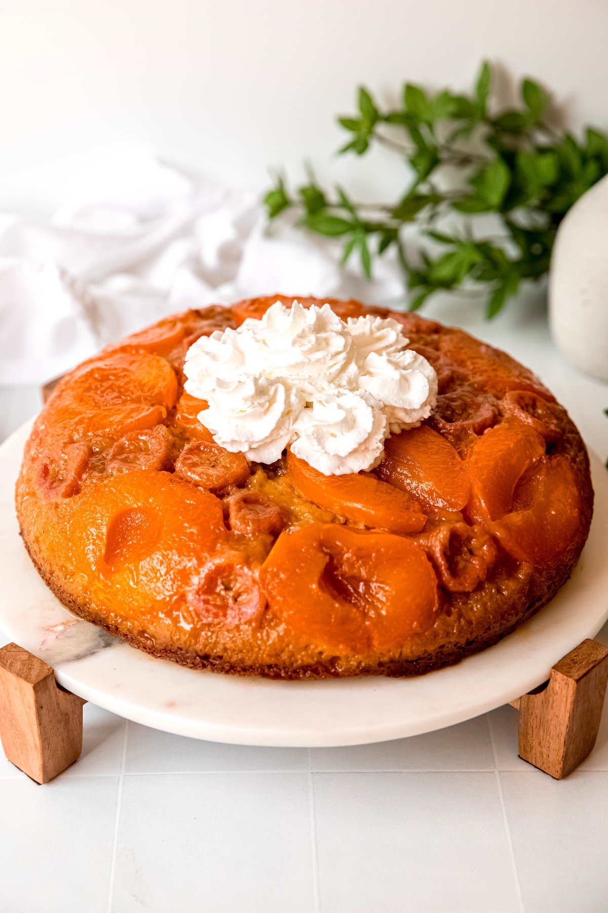 almond banana apricot upside down cake on a white marble cake stand with wooden legs topped with several swirls of whipped cream on a white table with a green houseplant.
