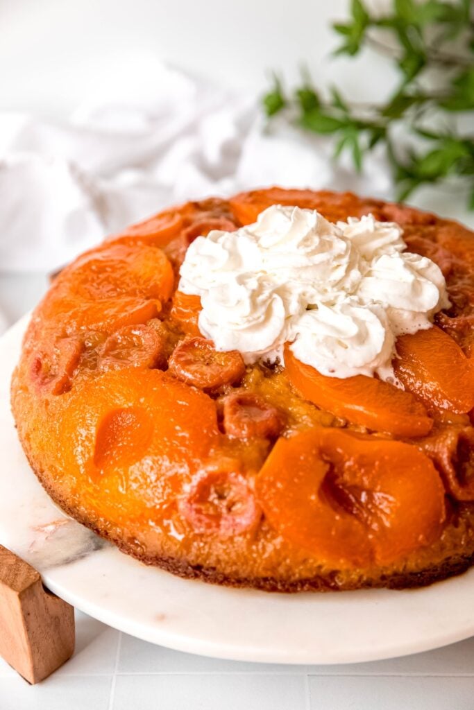 left ¾ of a cake mix banana apricot upside down cake topped with swirls of whipped cream on a white marble cake stand.