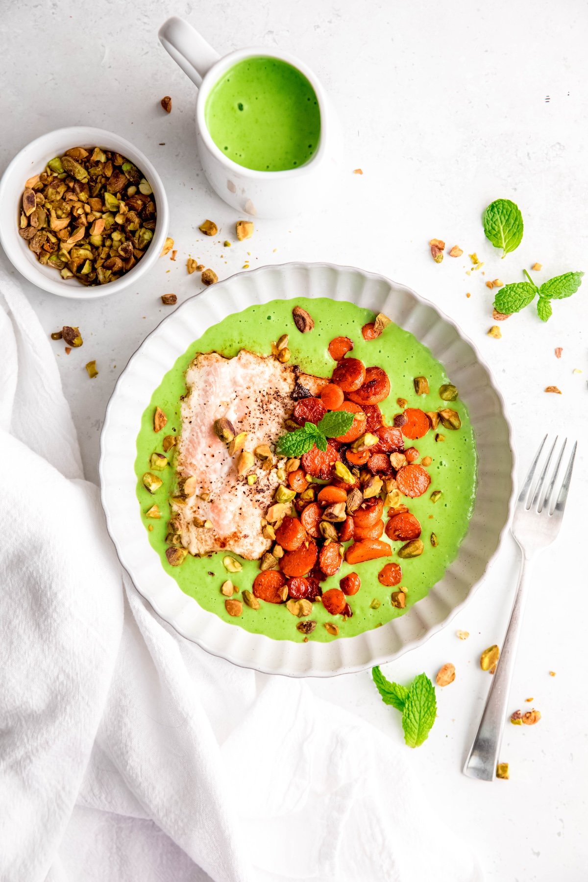butter basted salmon on a bed of mint pea puree with caramelized carrot medallions and pistachios in a white pasta bowl on a white table with a ramekin of extra pistachios, a small creamer dish filled with extra pea puree, and fresh mint leaves.