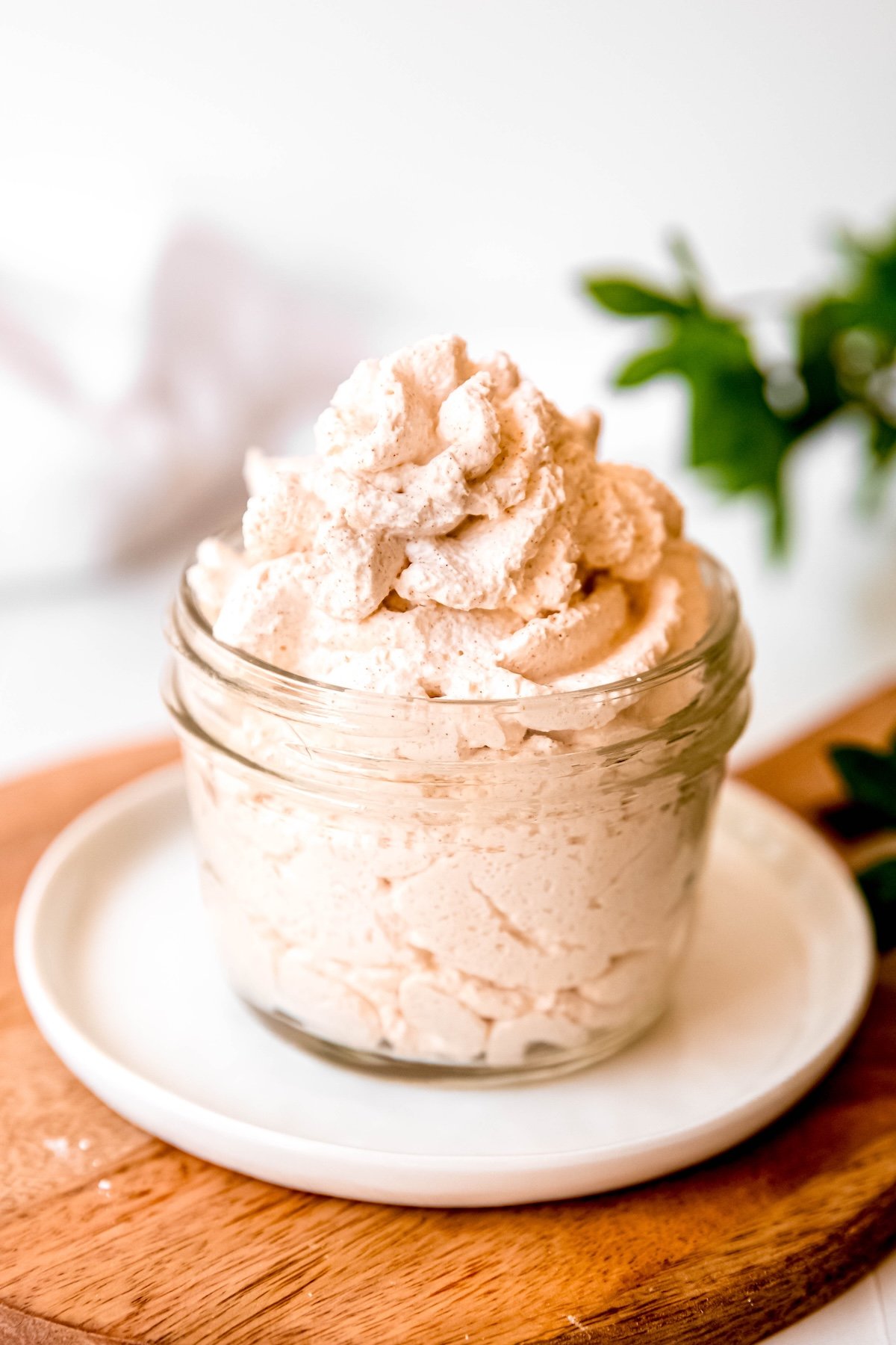 cinnamon whipped cream in a small mason jar on a white plate on a wooden serving board; it's light tan, and there's a green houseplant blurred in the background.