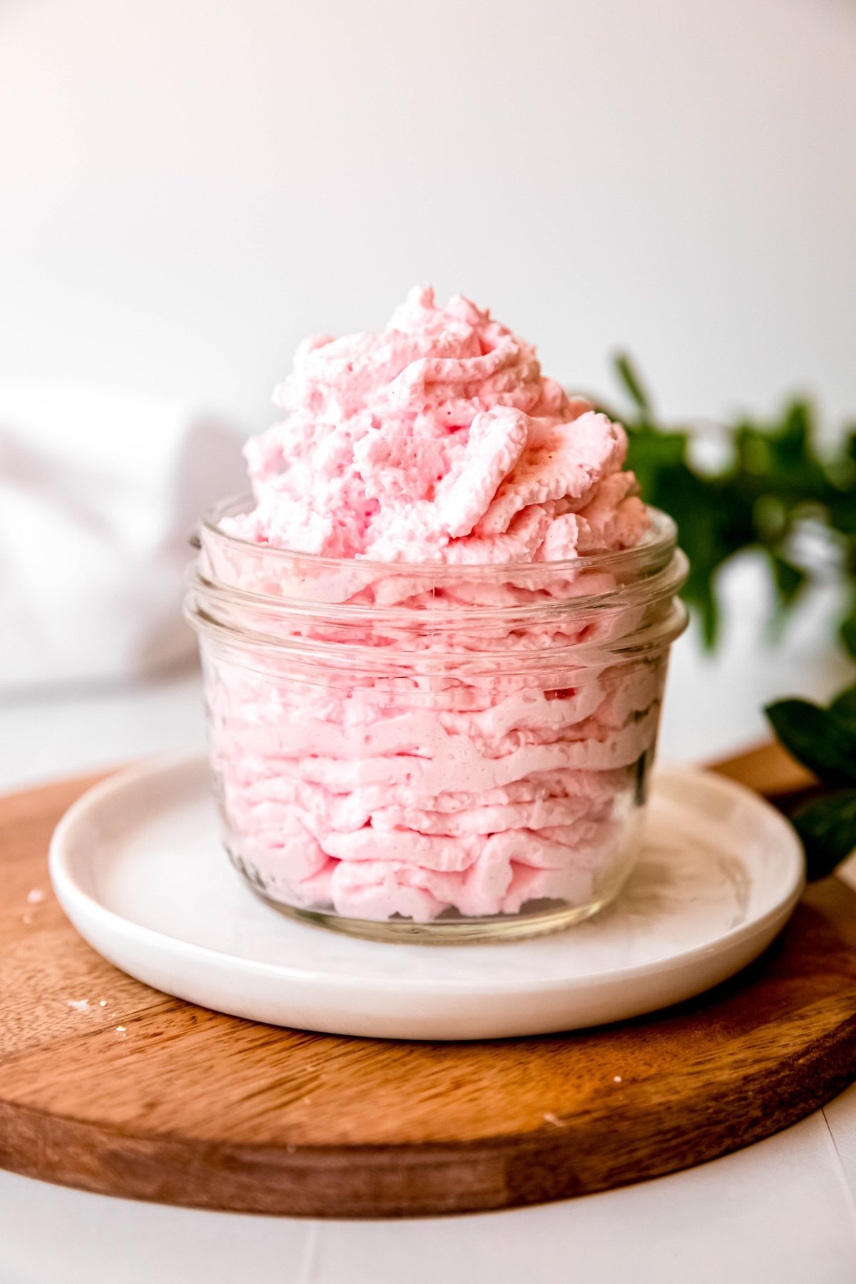 pale pink strawberry whipped cream in a small clear glass jar on a white plate on a wooden serving tray on a white table with a green houseplant blurred out behind.