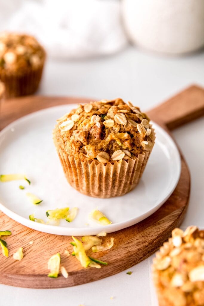 egg free zucchini muffins on a white table with the middle one on a white plate on top of a wooden serving tray with zucchini shreds.