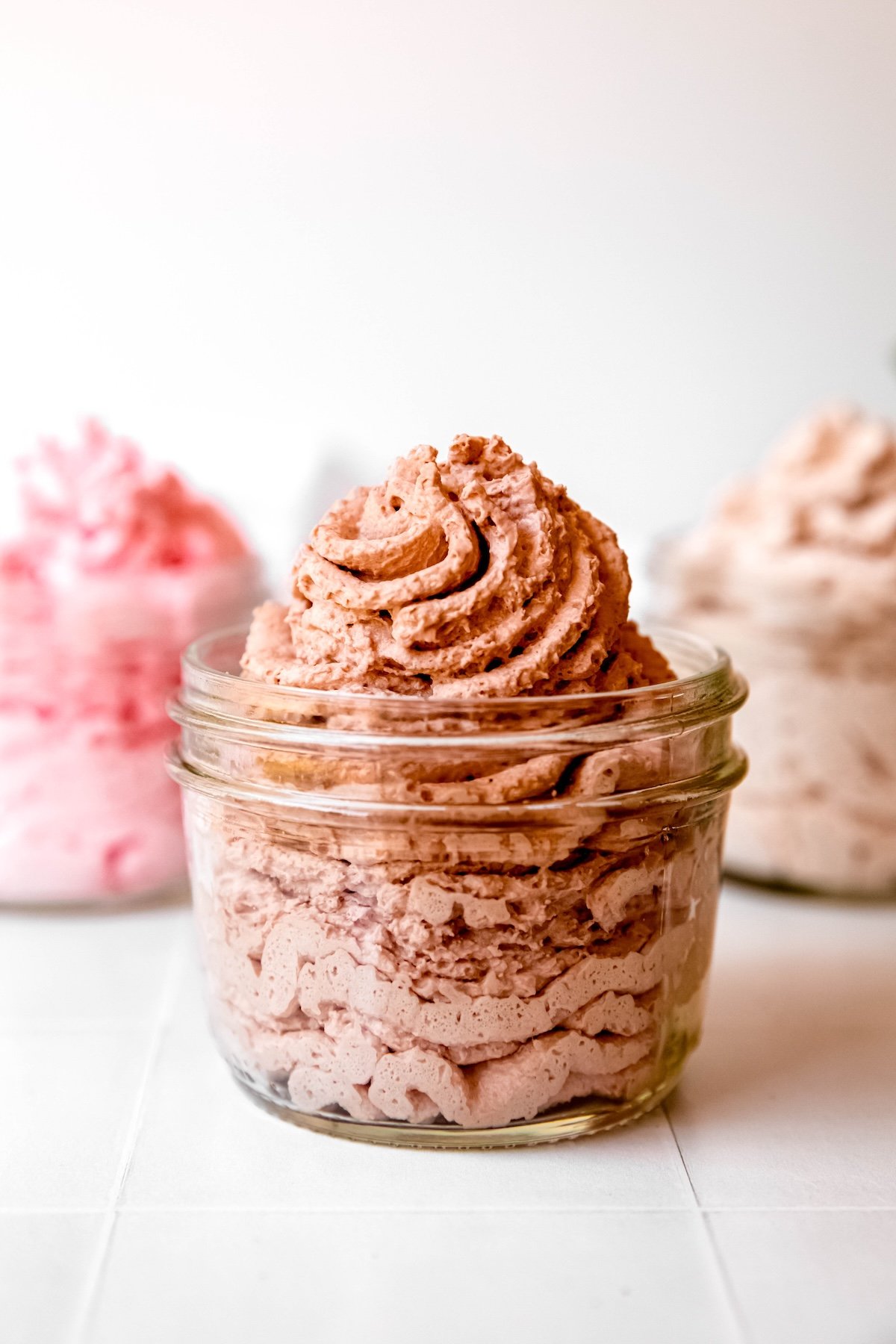 3 flavors of whipped cream dispenser recipes in clear jars on a table; chocolate is in the front, strawberry and cinnamon in the back.