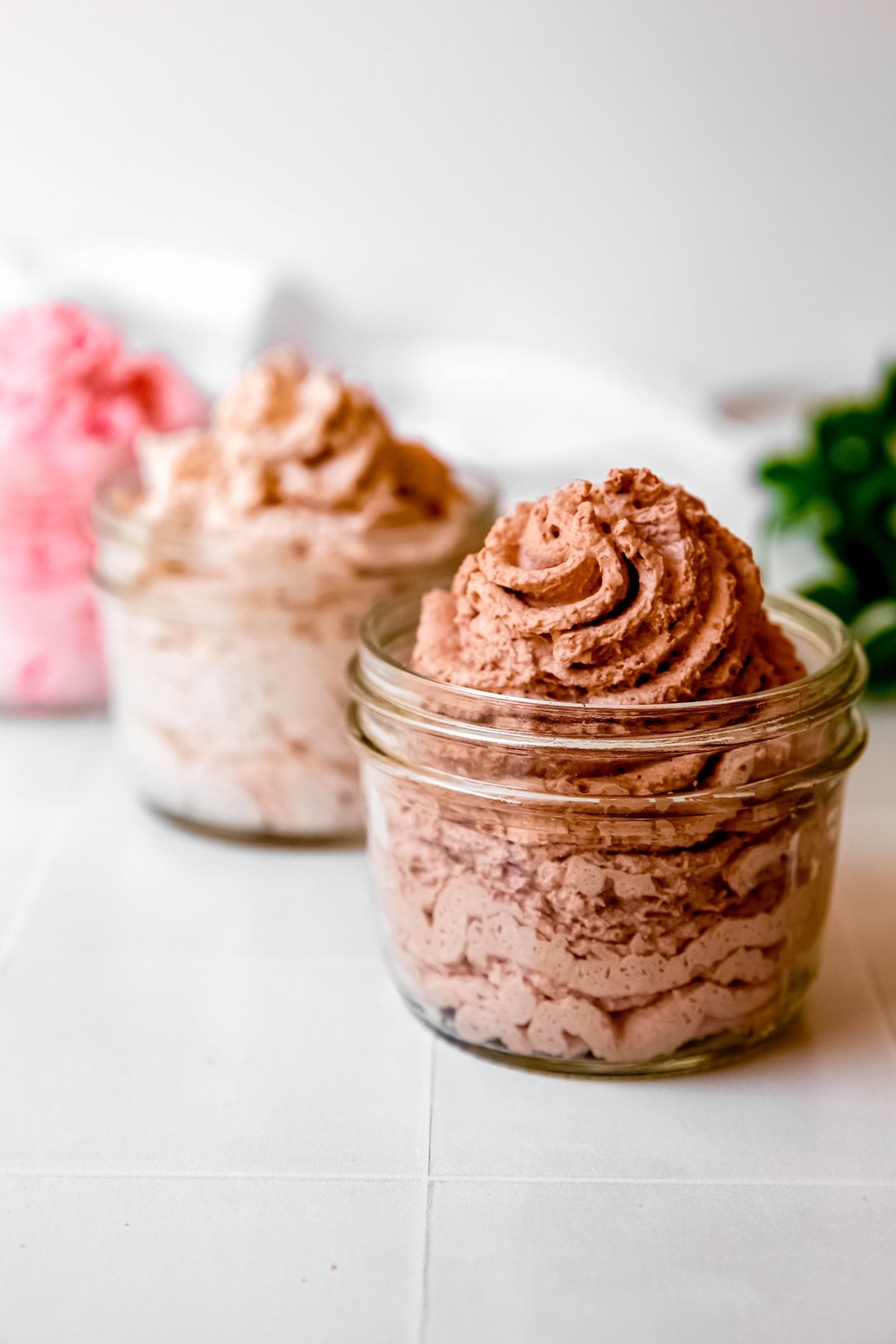 three small mason jars filled with chocolate whipped cream, cinnamon whipped cream, and strawberry whipped cream from a whipped cream dispenser lined up diagonally on a white table; the chocolate is in focus while the other two flavors are blurred out.