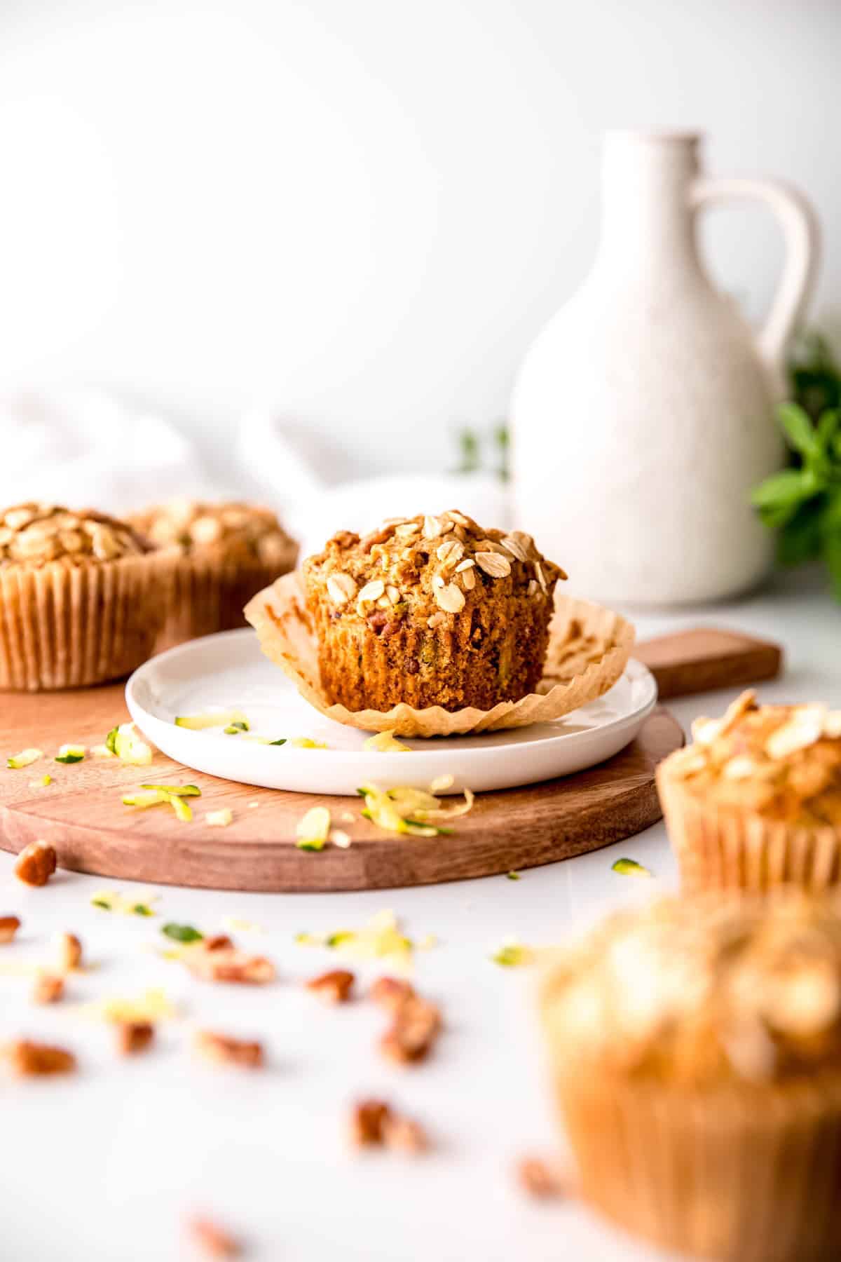 table of no egg oatmeal zucchini muffins with the center one on a white plate with the muffin liner pulled most of the way off; there's chopped nuts and grated zucchini sprinkled around.