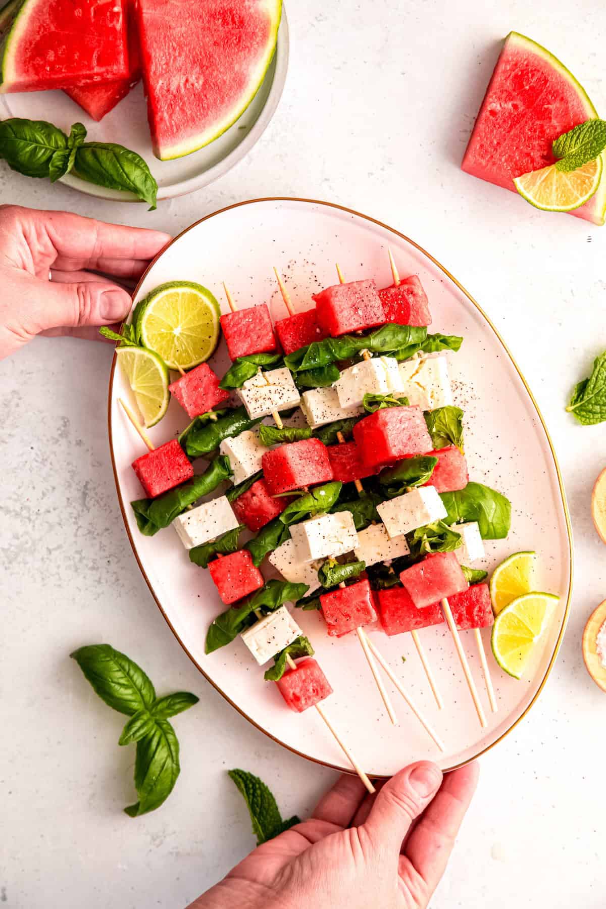 hands setting an oval white serving platter filled with savory watermelon skewers with feta and herbs down on a white table.