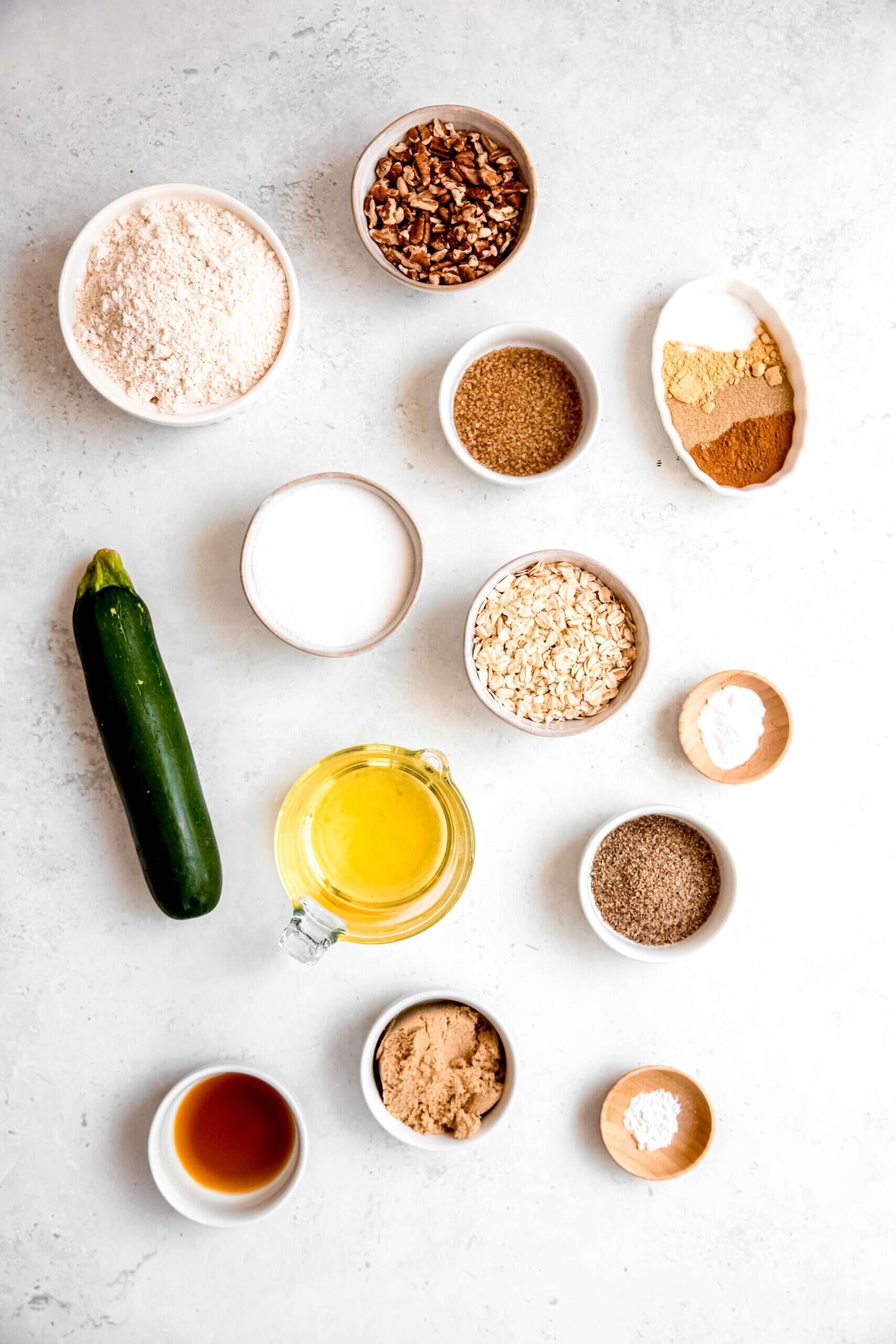 ingredients needed to make vegan whole wheat oatmeal zucchini muffins recipe measured out into bowls on a white table.