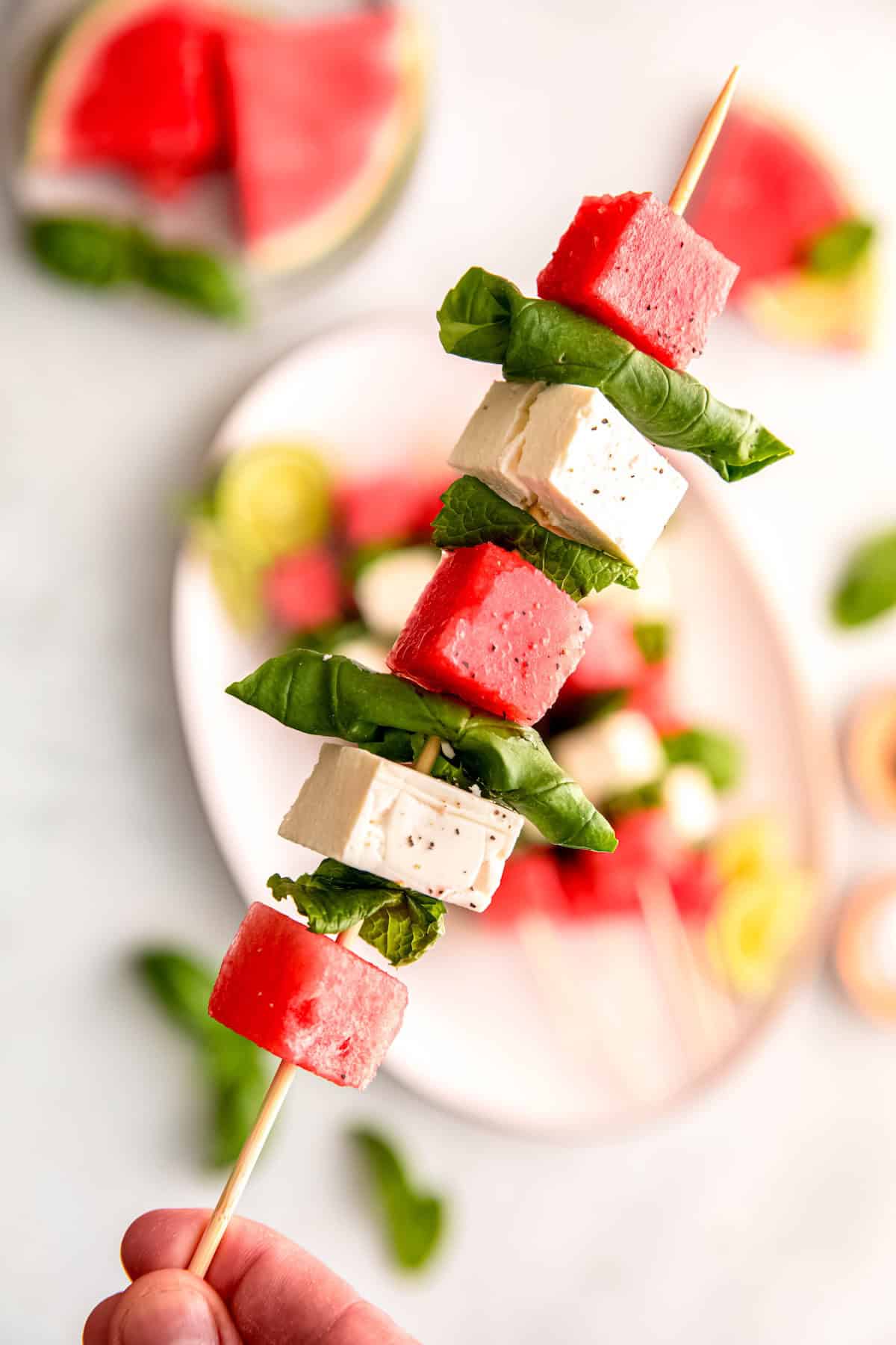 hand holding a single watermelon feta basil mint skewer above the rest of the platter blurred out beneath; you can see the shininess of olive oil and flakes of salt and pepper.