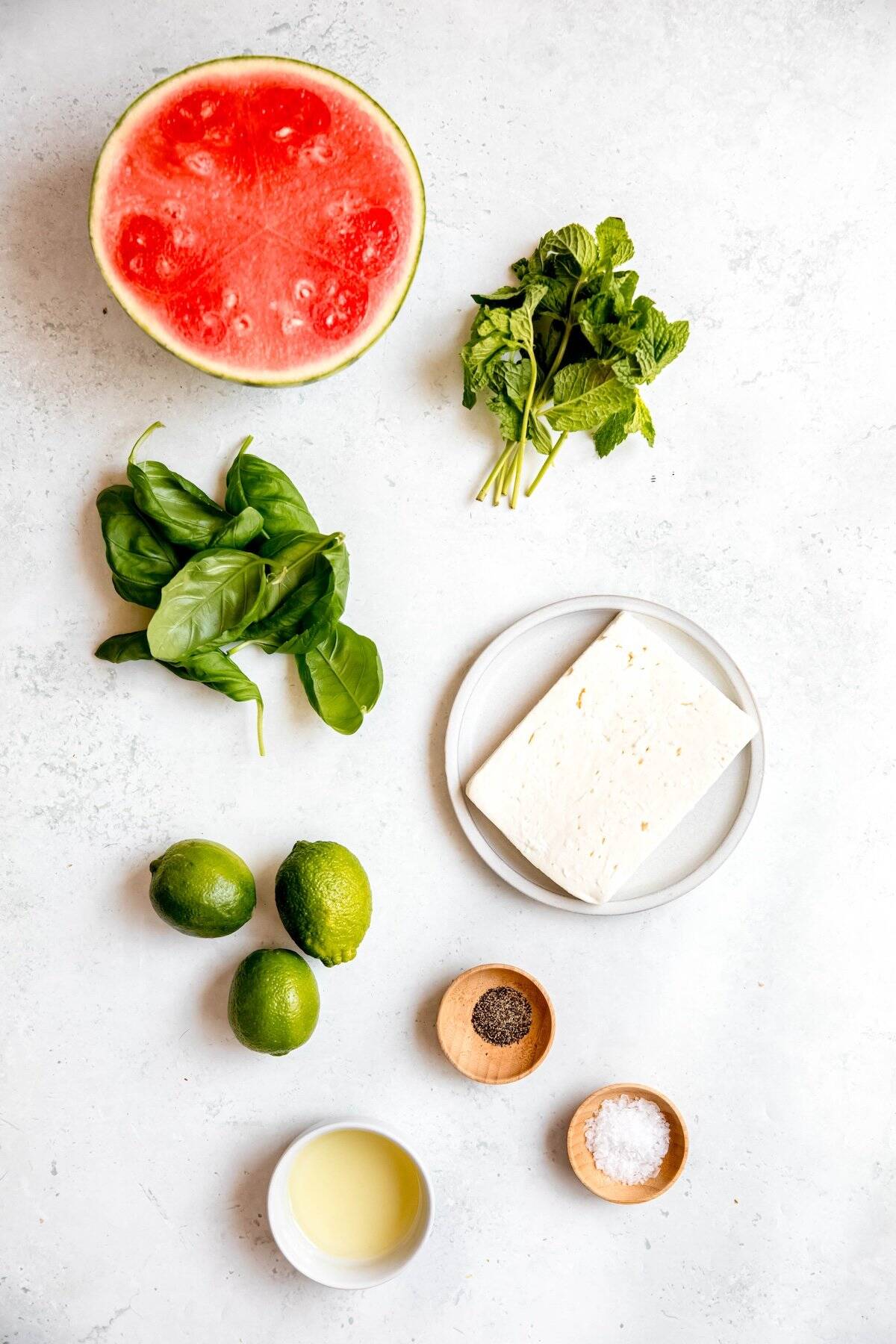 half a watermelon, a handful of fresh basil leaves, a bunch of fresh mint, a block of feta cheese, 3 fresh limes, olive oil, salt, and pepper measured out for making watermelon feta skewers.